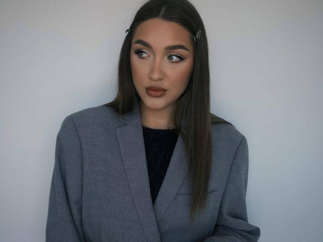 HeePickman has long brown hair, dark eyebrows, and brown lipstick. She's wearing a gray blazer over a black top, looking to the side with a calm expression.