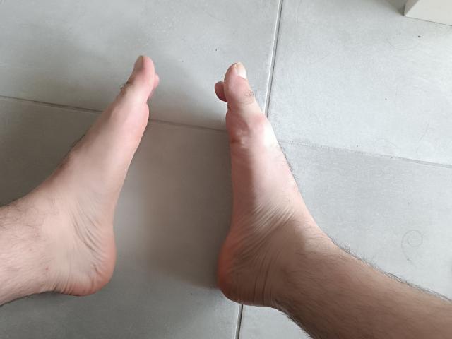 OrianaAndFriends' feet are on a tile floor, showing soles with visible creases and slight hair on the lower legs, toes pointing in different directions.