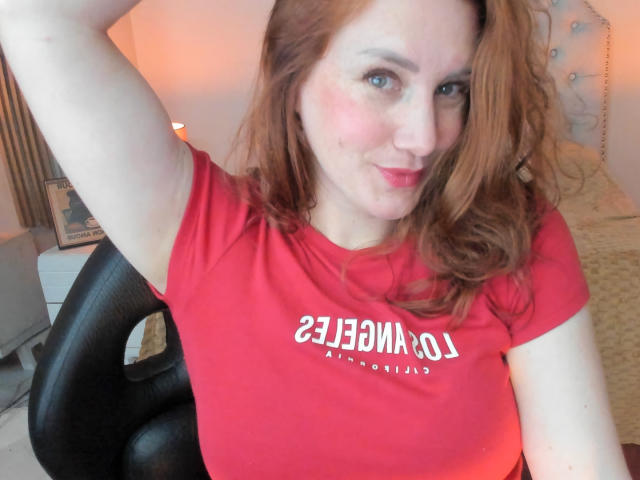KatyaRogersX with red hair, pale skin, and blue eyes, wearing a red shirt, raising her right arm, and smiling slightly, showing a small red mark on her cheek.