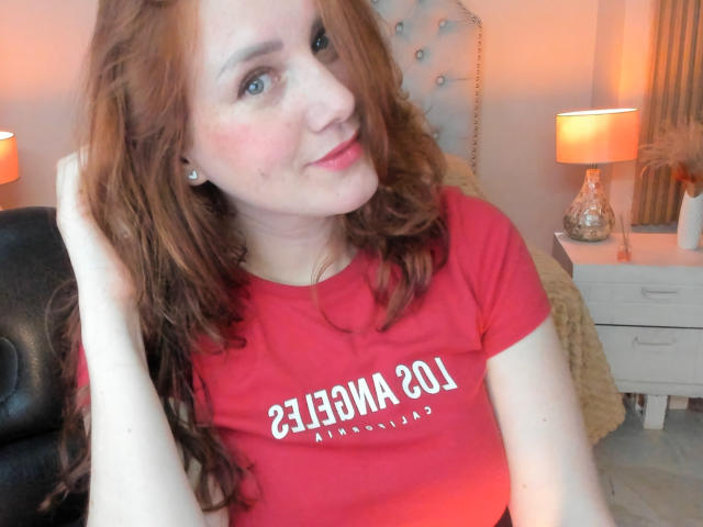 KatyaRogersX with red hair, wearing a red shirt, has a cute smile, blue eyes, and small tits, sitting in a chair, hand on head, in a softly lit room.