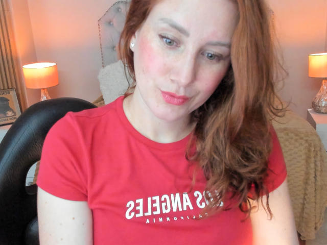 KatyaRogersX with red hair, red shirt, pale skin, slight blush, pink lips, sitting in chair, looking at camera, subtle smile, small nose, earrings, soft lighting, bedroom background.