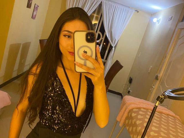CelesteFontaine with long brown hair, wearing a black sequined top with deep cleavage, holding a phone, taking a mirror selfie, showing her boobs and red lipstick.