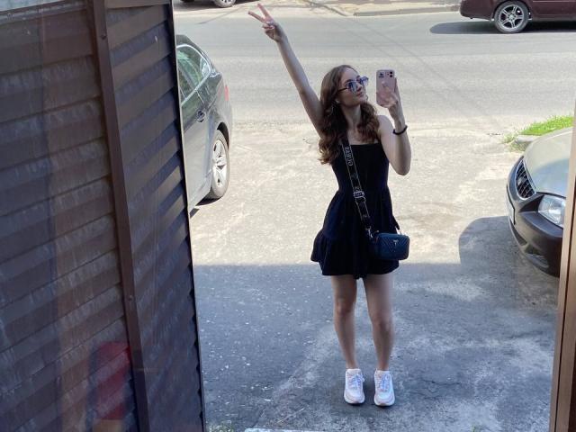 Gracie, en janvier, avec des cheveux bruns ondulés, portant une robe noire, des baskets blanches et un sac bandoulière bleu, faisant un signe de la paix, tenant un téléphone, debout dans une allée ensoleillée..