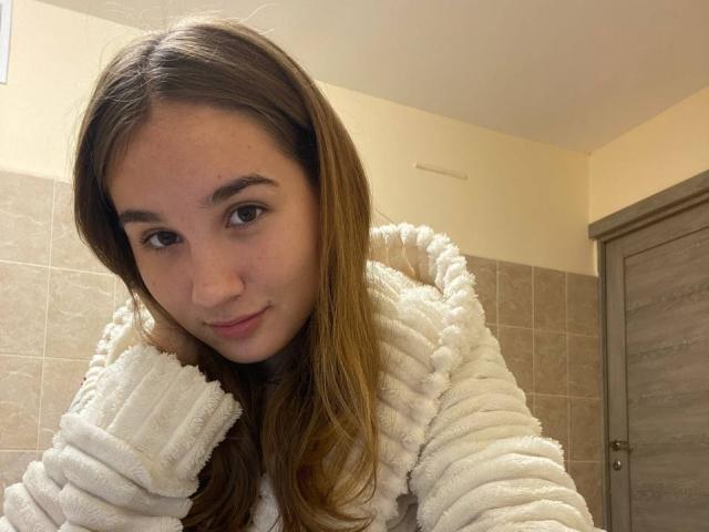 Gracie, im Januar geboren, mit langen braunen Haaren, trägt eine flauschige weiße Kapuzenjacke, hat ein süßes Lächeln und lehnt sich vor, wobei sie eine zarte Nase und weiche, braune Augen zeigt..