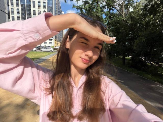 Gracie, en enero, con cabello castaño largo, camisa rosa, ojos entrecerrados, con la mano protegiéndose del sol, sonriendo, de pie en la acera, día soleado, árboles verdes al fondo, cielo azul claro..