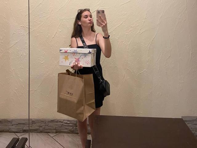 GracieJanuary está de pie frente a un espejo, tomándose una foto, vestida con un vestido negro, sosteniendo bolsas marrones y una caja de regalo colorida, con su trasero ligeramente visible, en una tienda..