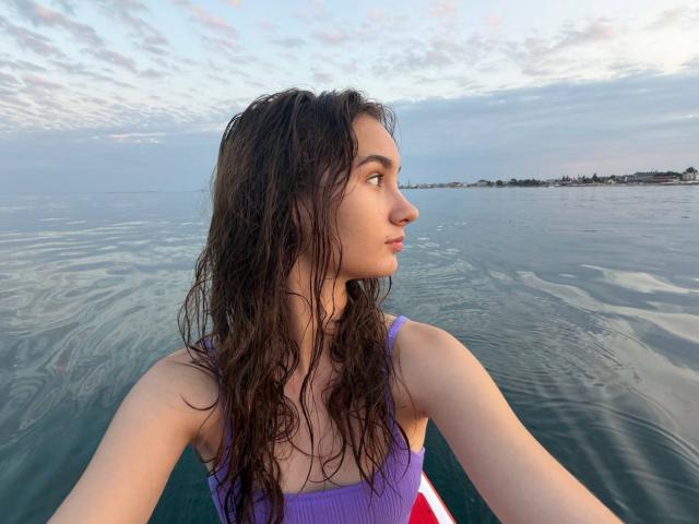 Gracie, en janvier, avec ses cheveux bruns foncés mouillés, porte un maillot de bain violet et regarde de côté depuis un bateau, montrant sa petite poitrine et son nez discret. L'eau est calme et le ciel est nuageux..