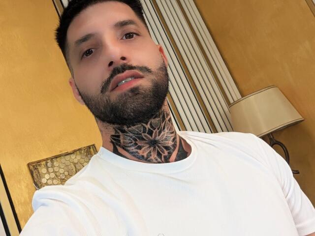 TommyDum with a beard and short black hair, wearing a white shirt, showing a detailed butterfly tattoo on his throat. His brown eyes are slightly open, lips parted.