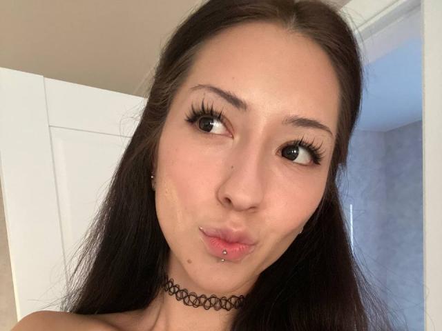 MarylnWerntz with long dark hair, pouty lips, small nose piercing, black choker, and thick eyelashes, giving a playful kissy face. Her eyes are slightly up, looking cute.