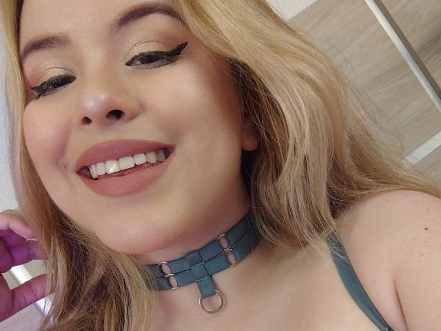 AshleyRossie with blonde hair, green eyeliner, pink lipstick, smiling, wearing a teal choker, visible teeth, holding a purple object, showing her tits in a green top.