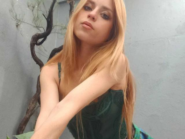 LyannaDifran with long blonde hair, green lace top, pensive expression, arms crossed, pale skin, small tits, green eyes, subtle makeup, standing in front of a gray wall.