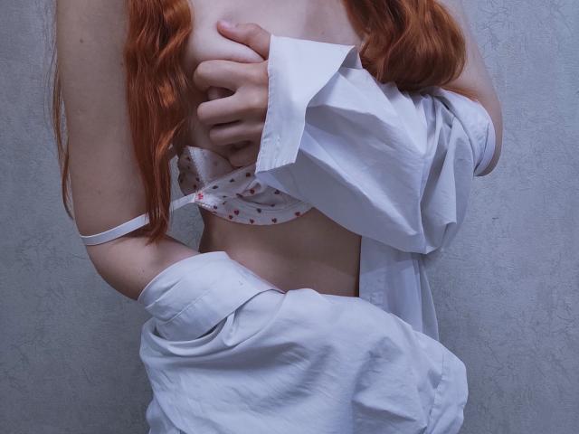 SheritaChieng with red hair, covering her tits with a white shirt, wearing a white bra with red hearts, and white pants, her hand on her chest, against a gray wall.