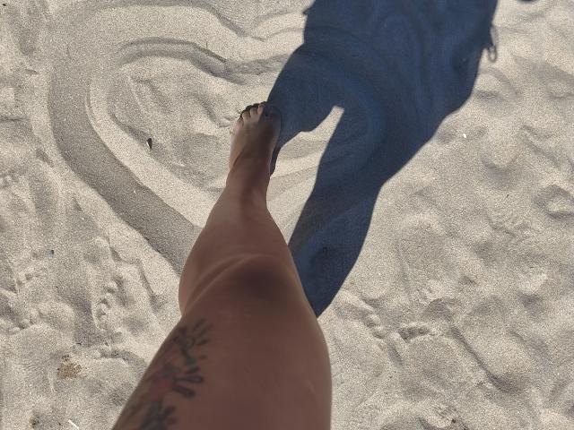 LilithGreenn's naked, tattooed thigh and foot in white sand, shadow of her legs and feet, visible blue shadow on sand, clear sunlight, small sand patterns.