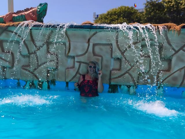LilithGreenn with her tits barely above water, wearing a red and black swimsuit, standing under a waterfall in a blue pool, with water spraying around her.