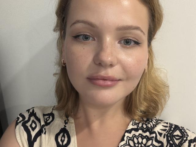 Carly Burross tem uma pele brilhante, com sardas, olhos azuis e cabelos loiros claros e ondulados. Ela usa delineador preto, pequenos brincos e uma blusa branca com estampas e detalhes pretos..
