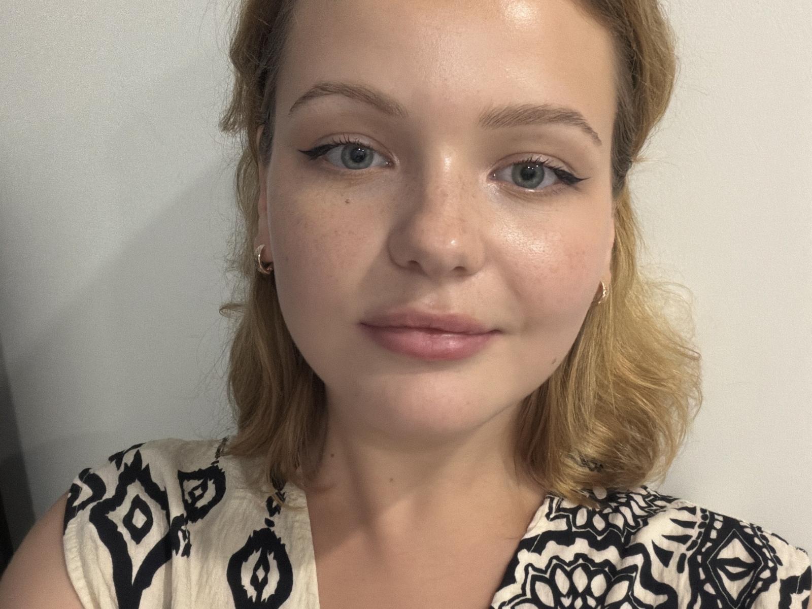 CarlyBurross has shiny, freckled skin, blue eyes, and light blonde, wavy hair. She wears black eyeliner, small earrings, and a patterned white top with black designs.