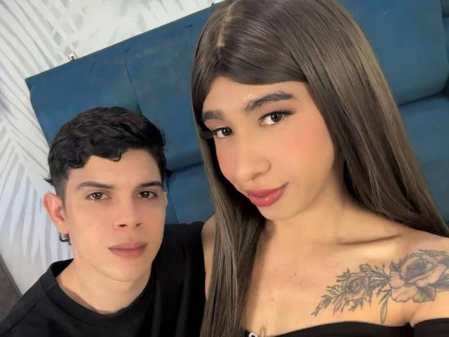 SecretOfTwo with long brown hair and pink lips, has a rose tattoo on her chest. She's next to a curly-haired boy in a black shirt, both smiling.