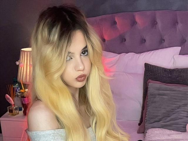 AmirraLovely, con lunghi capelli biondi, labbra piene e una maglietta a spalle scoperte, seduta su un letto con una testiera viola imbottita, che guarda di lato, in un ambiente da camera da letto..