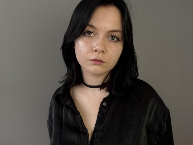 Elfrieda Leever tem pele clara, cabelo preto na altura dos ombros, olhos verdes, um colar preto e veste uma camisa preta com botões. Sua expressão é neutra e ligeiramente séria..