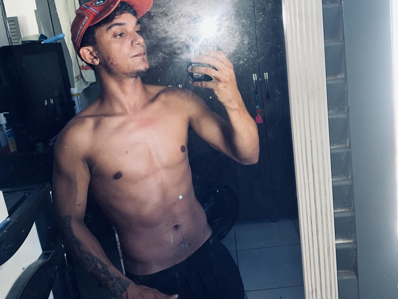 MatewsDias, senza camicia, con un cappello rosso, che tiene un telefono in un selfie allo specchio, mostrando un uomo atletico, tatuato, magro, con la pelle abbronzata, senza seno, circonciso, con il pene flaccido, che indossa pantaloni neri, muscoloso, attraente..
