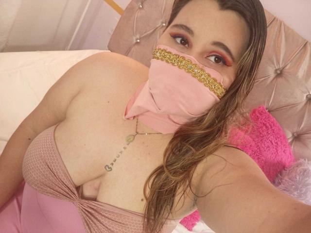 SalmaAzaman with wet brown hair, pink mask with gold trim, pink dress showing big boobs, tattoo on chest, pink pillow, and pink headboard in bedroom selfie.