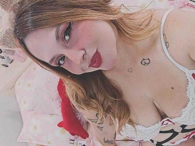 PauleetteRoux with red lipstick, nose ring, and tattoos, looking at camera, wearing white lace bra, showing her tits, blonde hair, pink headband, lying on bed.