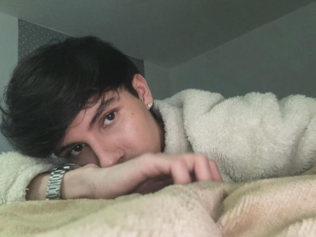 Angelitocute mit dunklem Haar, braunen Augen und einem Nasenring liegt auf einer beigen Decke, trägt ein flauschiges weißes Hemd und eine silberne Uhr und blickt mit einem entspannten Ausdruck nach oben..