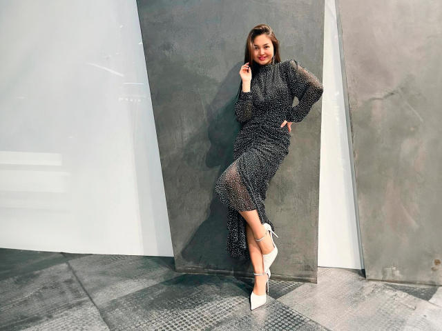 KetrinMagicx está de pie en una habitación gris, vestida con un vestido negro transparente de manga larga, tacones blancos y una sonrisa segura. Su mano descansa sobre su cadera, con las piernas cruzadas..