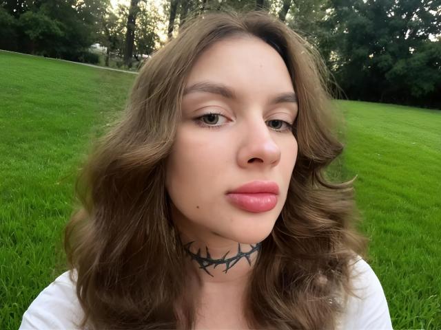 AlisTris tiene el pelo castaño ondulado, lápiz labial rosa y un collar negro con espinas. Está en un campo lleno de hierba, con árboles al fondo, y parece un poco aburrida..