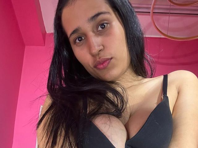 KinkyIris has dark hair, brown eyes, pink lips, and a black bra that shows her big tits. She's standing in a pink room with a ceiling fan.
