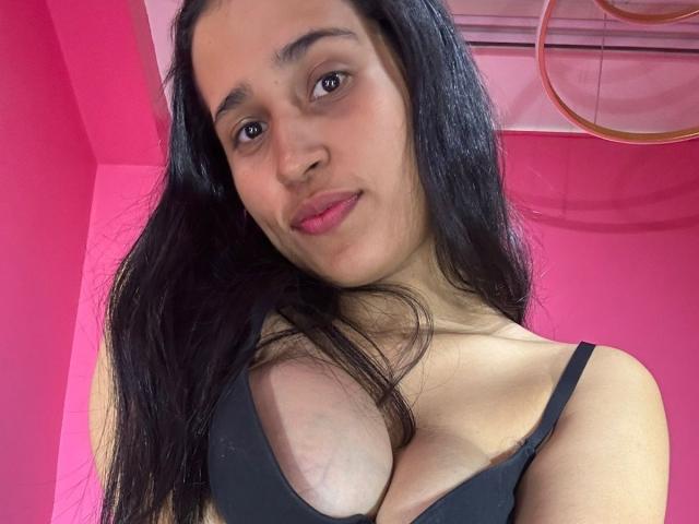 KinkyIris, com cabelos pretos longos, olhos castanhos e lábios rosados, vestindo um sutiã preto que mostra seus seios grandes, está em pé em frente a uma parede rosa. Ela está sorrindo suavemente para a câmera..