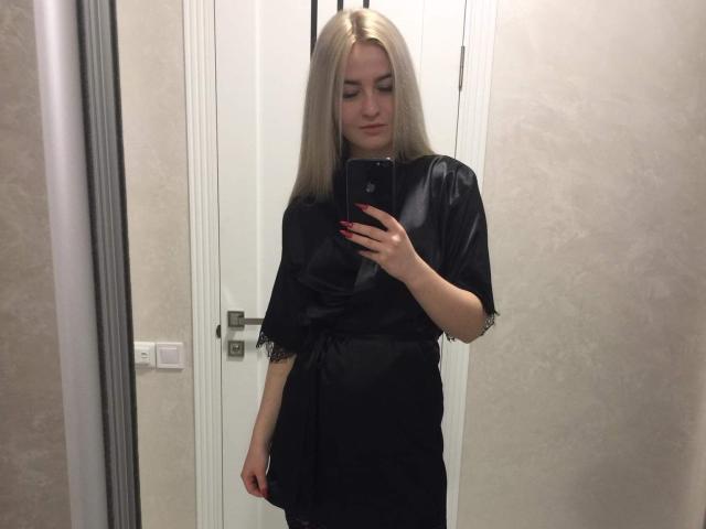 JulieBlaze with long blonde hair, wearing a black satin robe, taking a mirror selfie with a black phone, showing her red nails and a subtle smile, in a room.