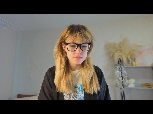 HortenciaScudder with blonde hair, black glasses, and a black shirt over a white tee, looking slightly down with a subtle smile. She's got cute tits and a relaxed vibe.