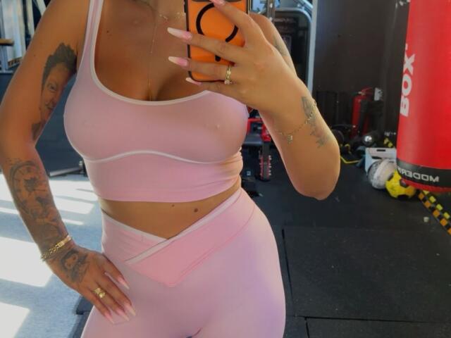 NicolSNaya with pink sports bra and matching pink booty shorts, showing her big tits and curvy ass, holding an orange phone, tattooed arm, in a gym.