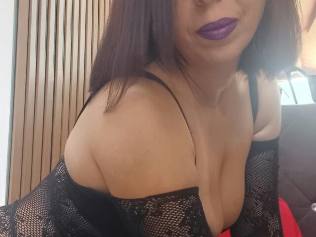 LauraSin with purple lipstick, black lace top, showing her tits, light skin, brown hair, and a slight smile, leaning forward. Her top is off-shoulder, exposing cleavage.
