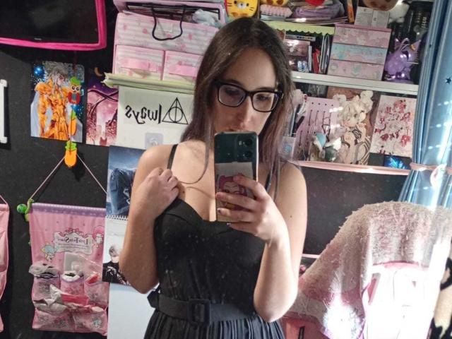 Rosabelle Jeannette, portant des lunettes, tenant un téléphone, vêtue d'une robe noire, montrant de petites seins, tenant une bretelle, debout dans une pièce avec des sacs roses et blancs, des jouets en peluche et un manteau rose duveteux..