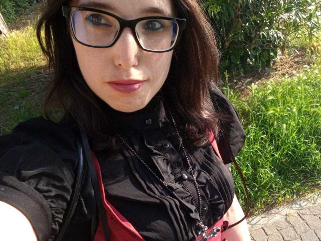 Rosabelle Jeannette mit Brille, dunklem Haar, schwarzem T-Shirt, roten Trägern, einer Halskette und pinkem Lippenstift, die auf einem Selfie im Freien ihre Brüste zeigt..