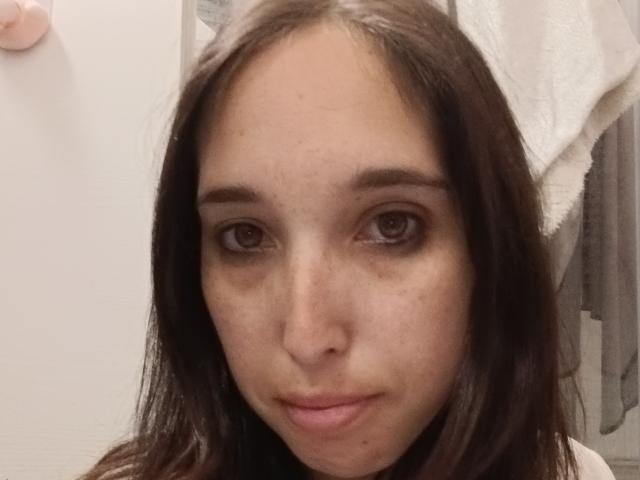 Rosabelles Gesicht mit braunen Augen, glattem, dunkelbraunen Haar, heller Haut und kleinen Sommersprossen, sie trägt ein weißes Handtuch im Haar, sieht leicht müde und verkniffen aus..