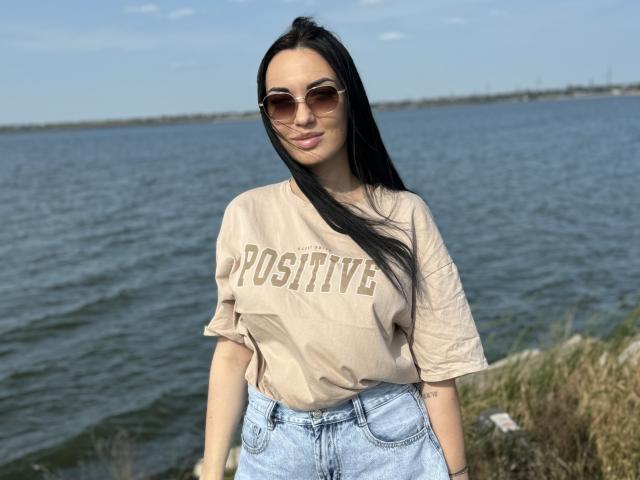 Xioni with long black hair, wearing a beige "Positive" t-shirt and blue jeans, standing by a lake, sporting brown aviator sunglasses, smiling softly.
