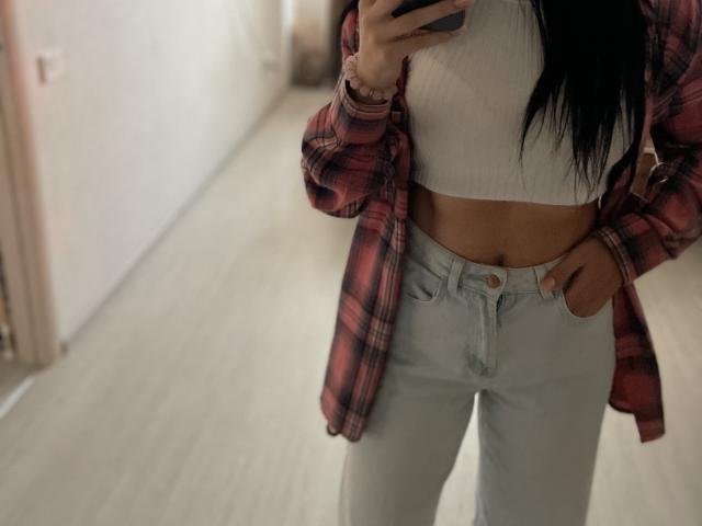 Xioni zeigte ihren Bauch, trug ein weißes, geripptes Crop-Top, ein rotes Karohemd und helgraue Jeans, ihre Hand steckte in der Hosentasche, sie hatte langes, schwarzes Haar und gebräunte Haut..