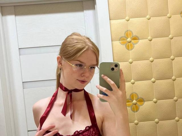 Shakia Ivester, com cabelo loiro, óculos, blusa vermelha de renda com alças finas e um colar vermelho de fita, segurando um telefone, cobrindo seus pequenos seios com uma mão, tirando uma selfie no espelho..