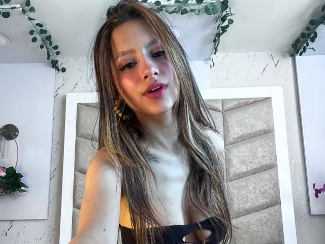 AfroditaWild has long brown hair, pink lipstick, and a cute face with small tits, wearing a black strapless top, standing in a room with green plant garland.