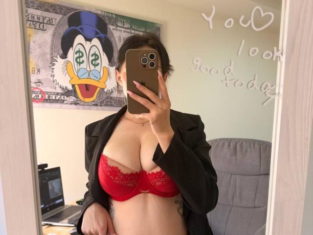 EmmaNoa with red lace bra, big boobs, black blazer, taking mirror selfie, holding gold phone, tattoo on side, laptop in background, text "You look good today" on mirror.