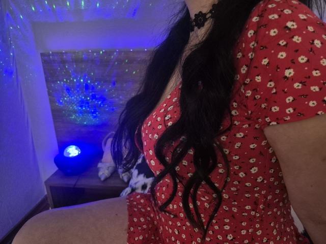 WenTina with long black hair, wearing a red floral dress, showing her boobs, sitting with a blue light in the background, and a black lace choker.