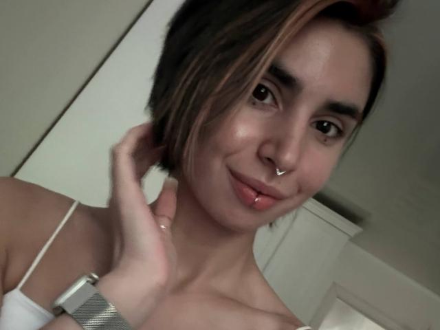 TameikaMacclellan with short brown hair, nose ring, and lip ring, smiling slightly, hand touching neck, wearing a white strap top, visible cleavage, soft lighting.