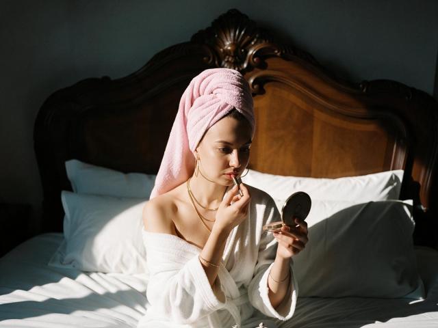 KamilaaXHot con un asciugamano rosa sulla testa, indossa un camice bianco, si sta truccando con il rossetto davanti a uno specchio, seduta su un letto con una testiera in legno, appare sensuale..