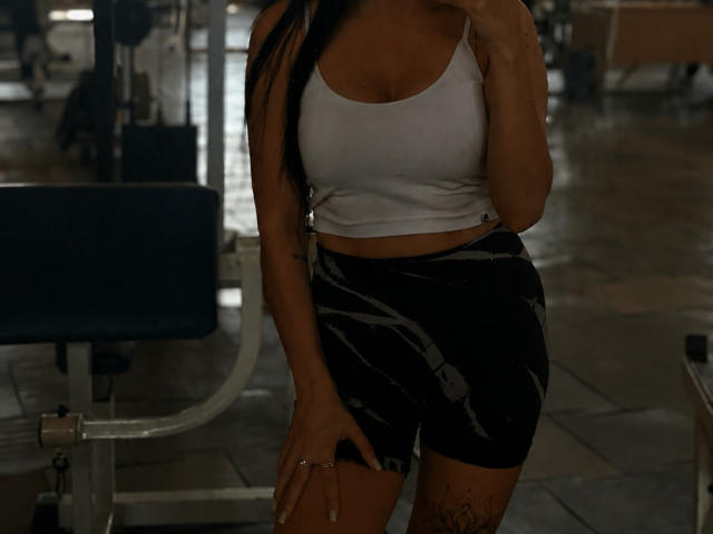 Xioni, con lunghi capelli neri, indossa un canottino bianco che mette in mostra il seno, pantaloncini neri marmorizzati, ha un tatuaggio sulla coscia destra, tiene in mano un telefono, è in piedi in una palestra poco illuminata, con un braccio che le copre la parte inferiore del viso..