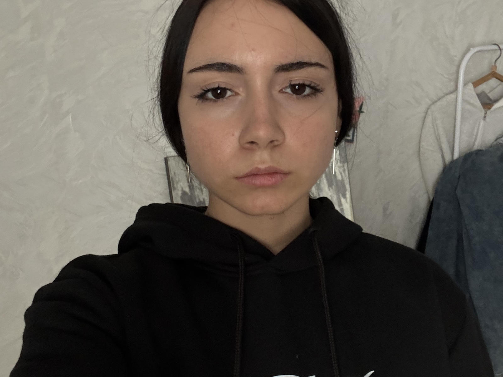 SusannValen, con piel pálida, cabello castaño oscuro y senos pequeños, lleva una sudadera negra, parece aburrida, tiene un maquillaje discreto y una pequeña cicatriz en la mejilla derecha..