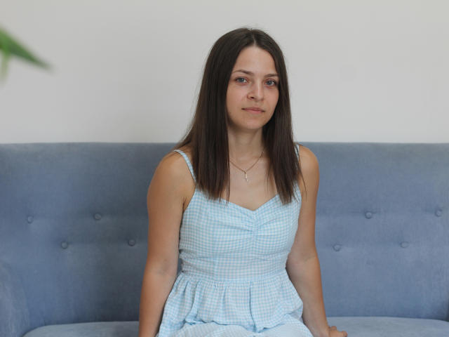 SaraUili tem cabelo castanho liso, pele clara e seios pequenos. Ela está vestindo um vestido azul xadrez, sentada em um sofá azul, com um sorriso discreto e um colar..