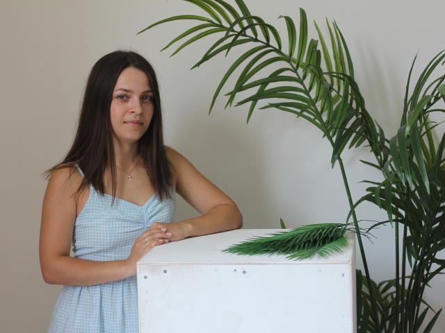 SaraUili with brown hair, light skin, wearing a blue check dress, standing by a white box with green palm leaves on top. She has small tits and a slight smile.